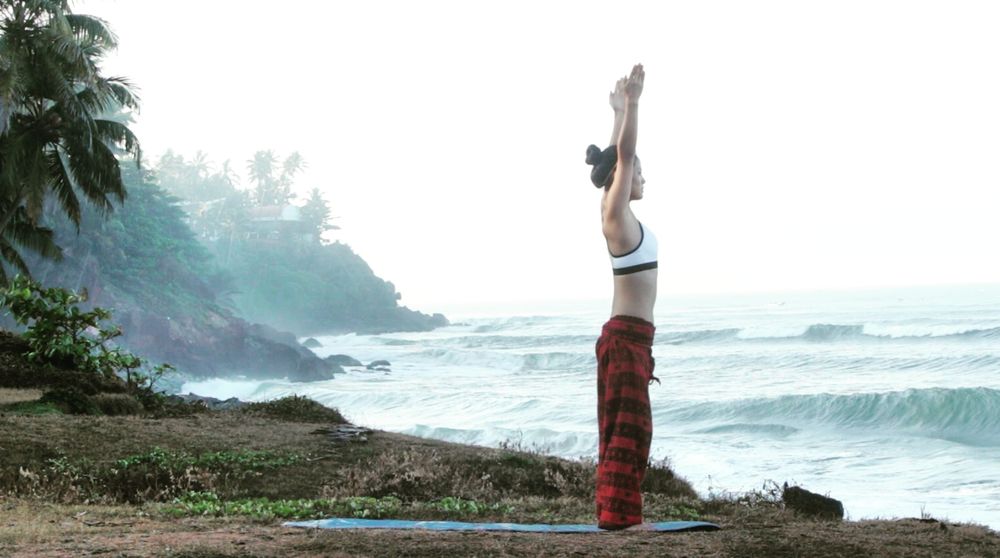 Anne Tana steht mit gestreckten Armen auf einer Yogamatte an einer nebligen Küste mit Palmen