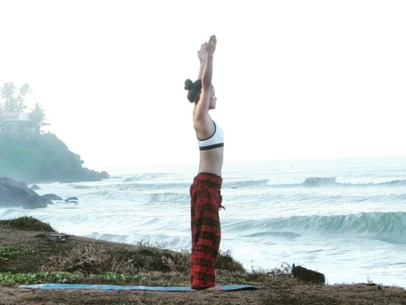 Anne Tana steht mit nach oben gestreckten Armen auf einer Yogamatte an einer nebligen Meeresküste