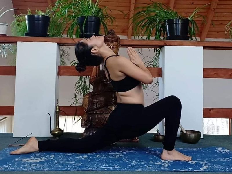 Anne Tana im tiefen Ausfallschritt mit gefalteten Händen in einem Yogaraum mit Holzdecke und Pflanzen